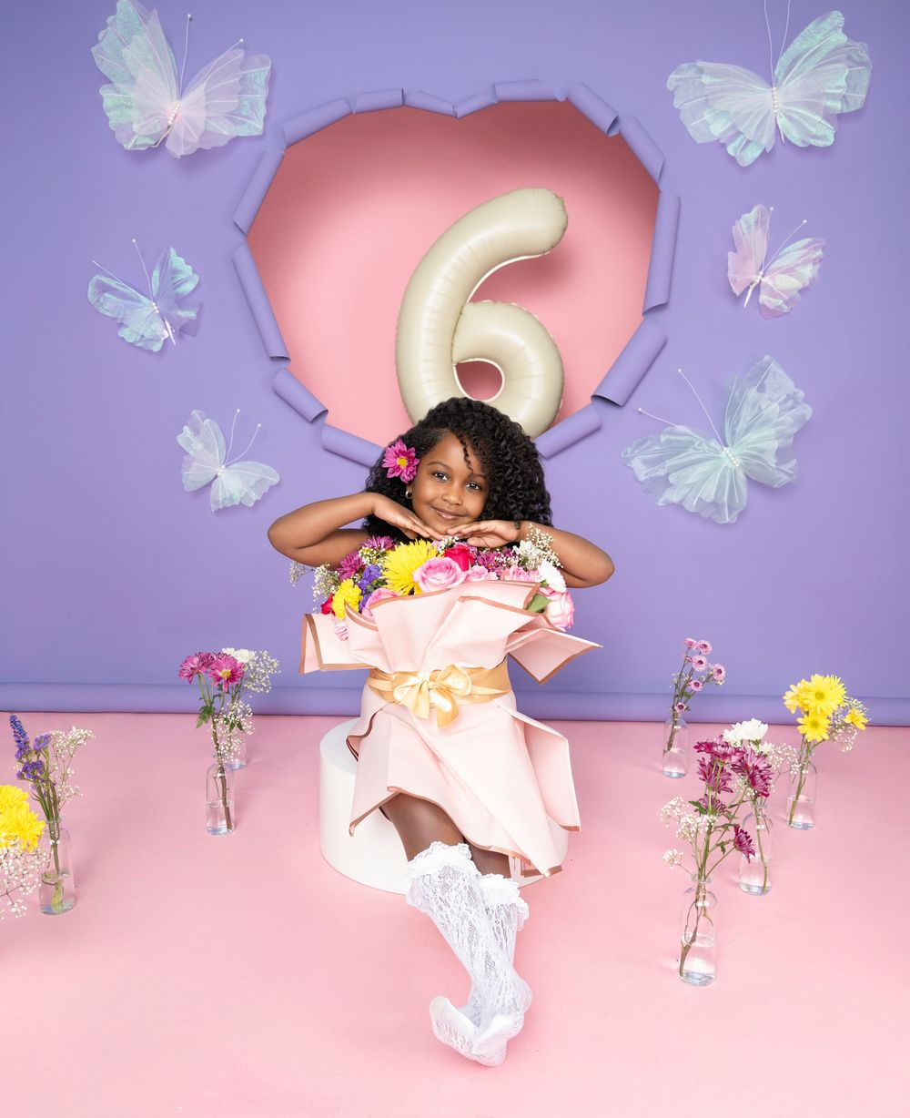 A child poses against a purple wall with a pink heart, number 6, and decorative butterflies for a birthday photoshoot.