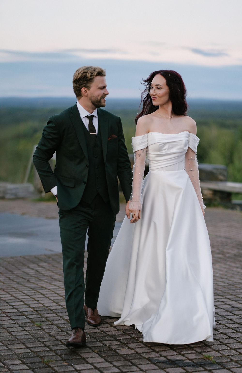 A couple in wedding attire walks together on a stone path with a scenic mountain landscape in the background.