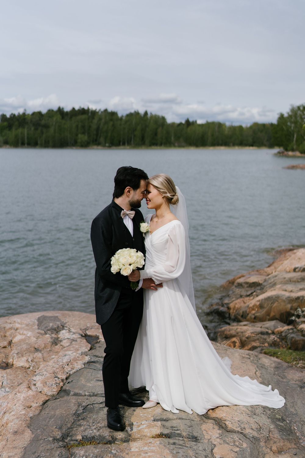 A couple shares an intimate moment on rocky shoreline with lake and forest backdrop.