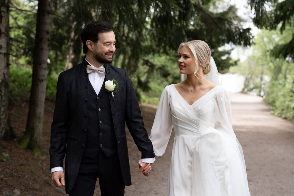 A happy couple walks hand in hand through a wooded path, she in an elegant white gown and he in a black tuxedo.