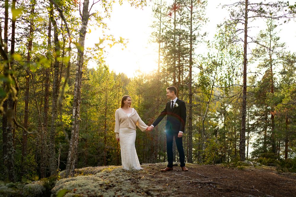 A couple in wedding attire holding hands while standing on a rocky overlook in a sunlit autumn forest.