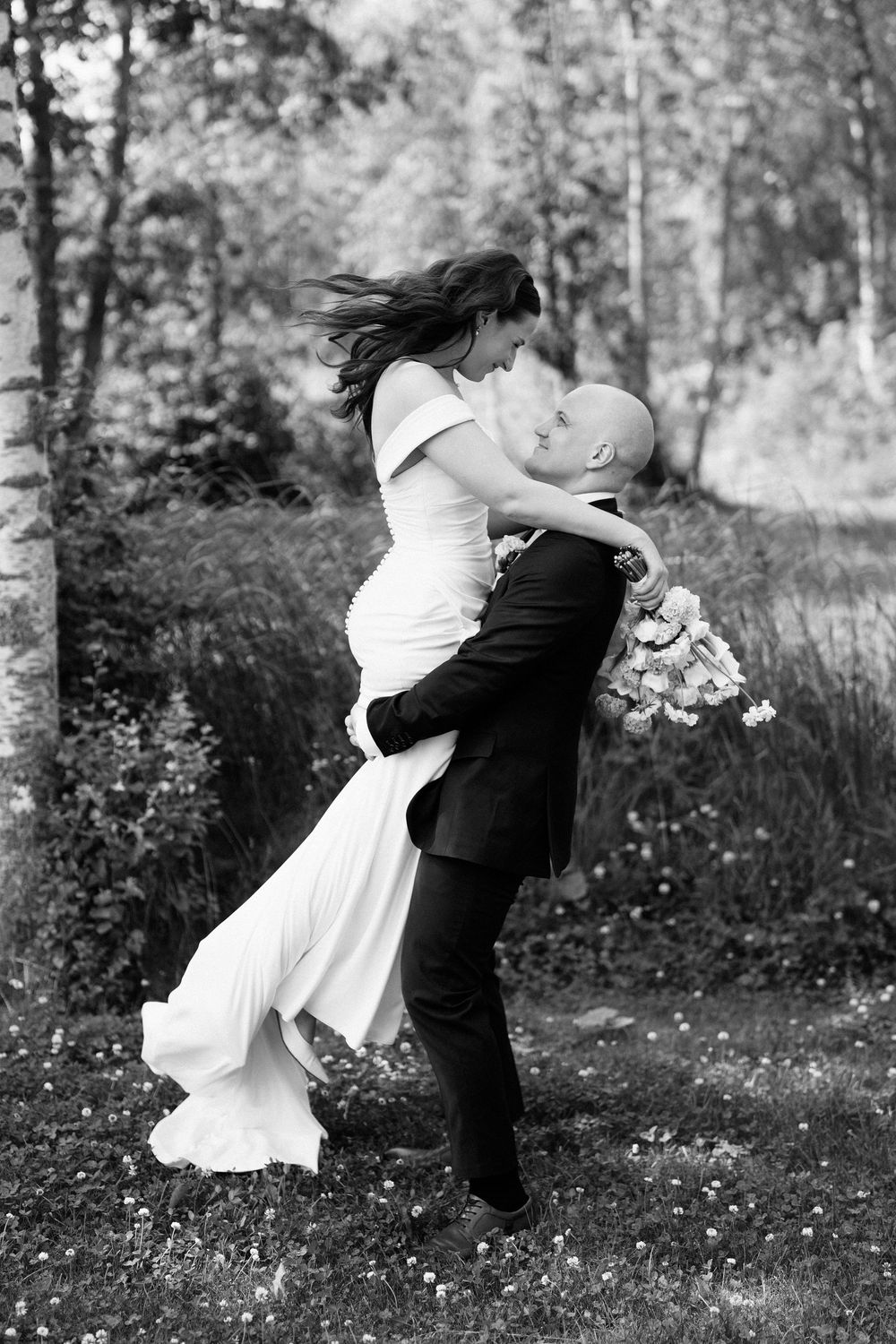 A romantic black and white wedding photo of a couple embracing outdoors in a wooded setting.