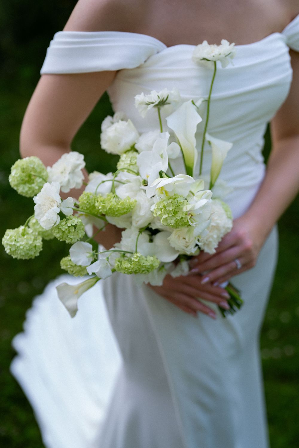 A close-up of a bridal bouquet featuring white and light green flowers against an off-shoulder wedding dress.
