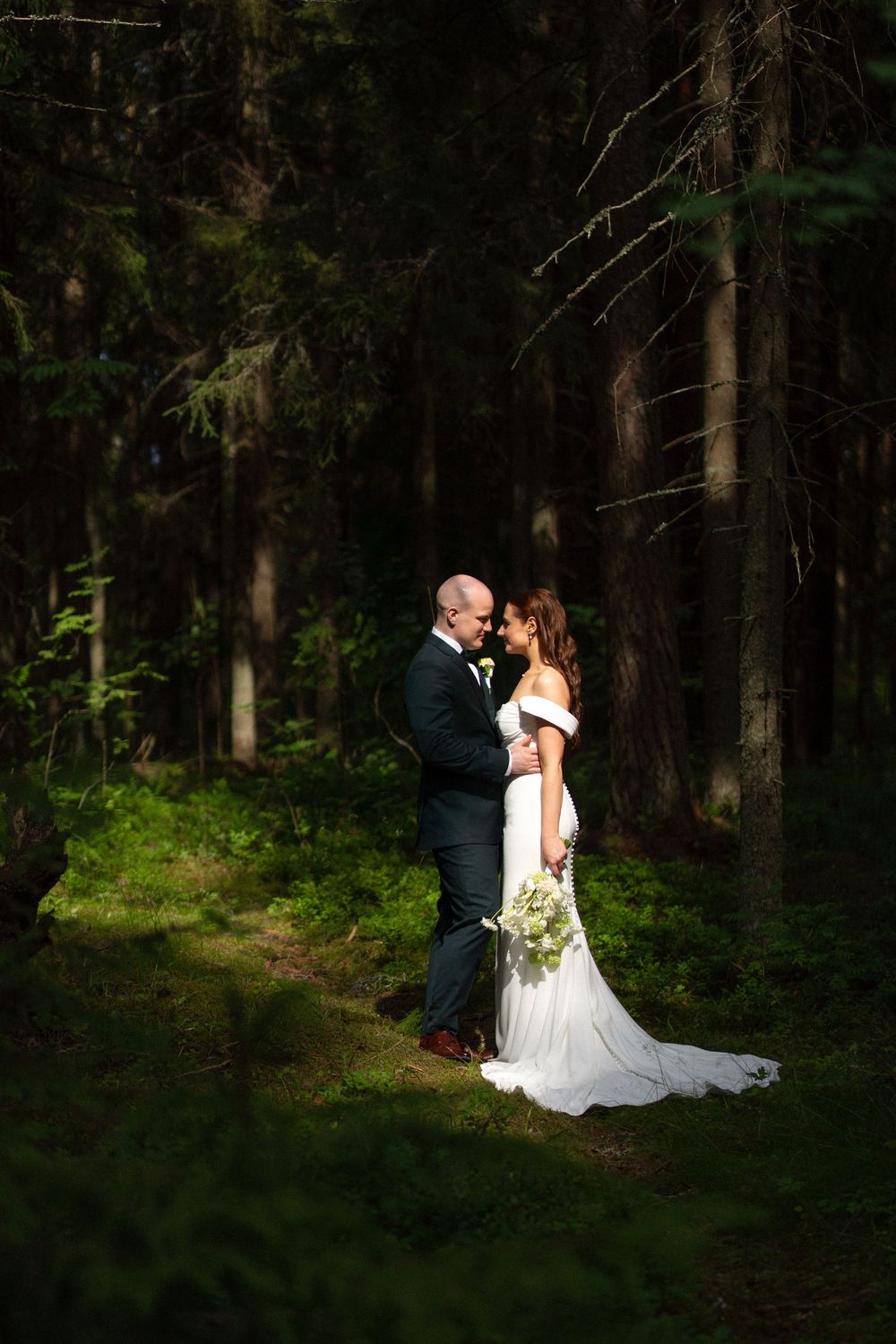 A romantic wedding couple shares intimate moments in a serene forest setting with dappled sunlight filtering through trees.