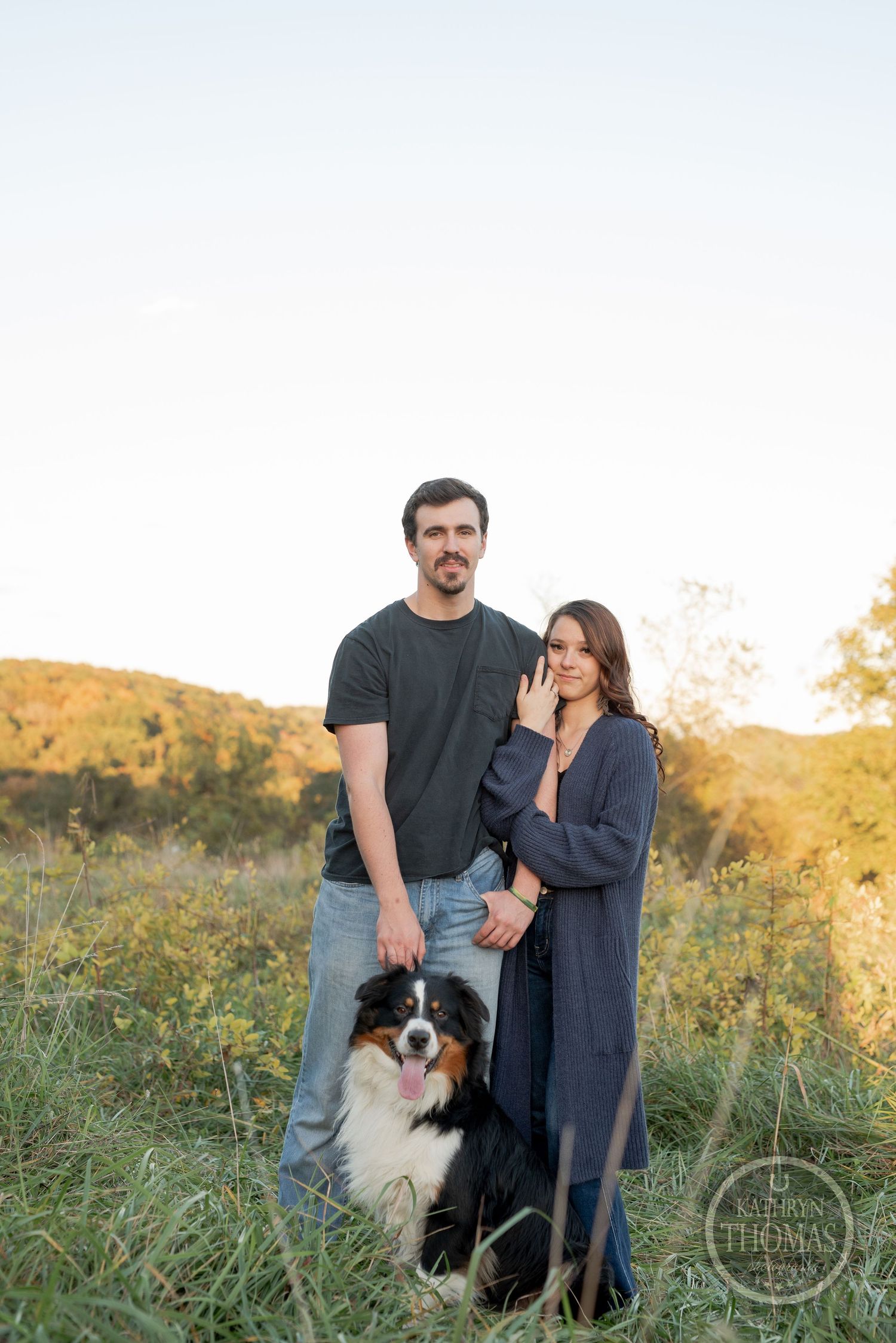 A couple poses with their Australian Shepherd dog in a scenic autumn field at sunset.