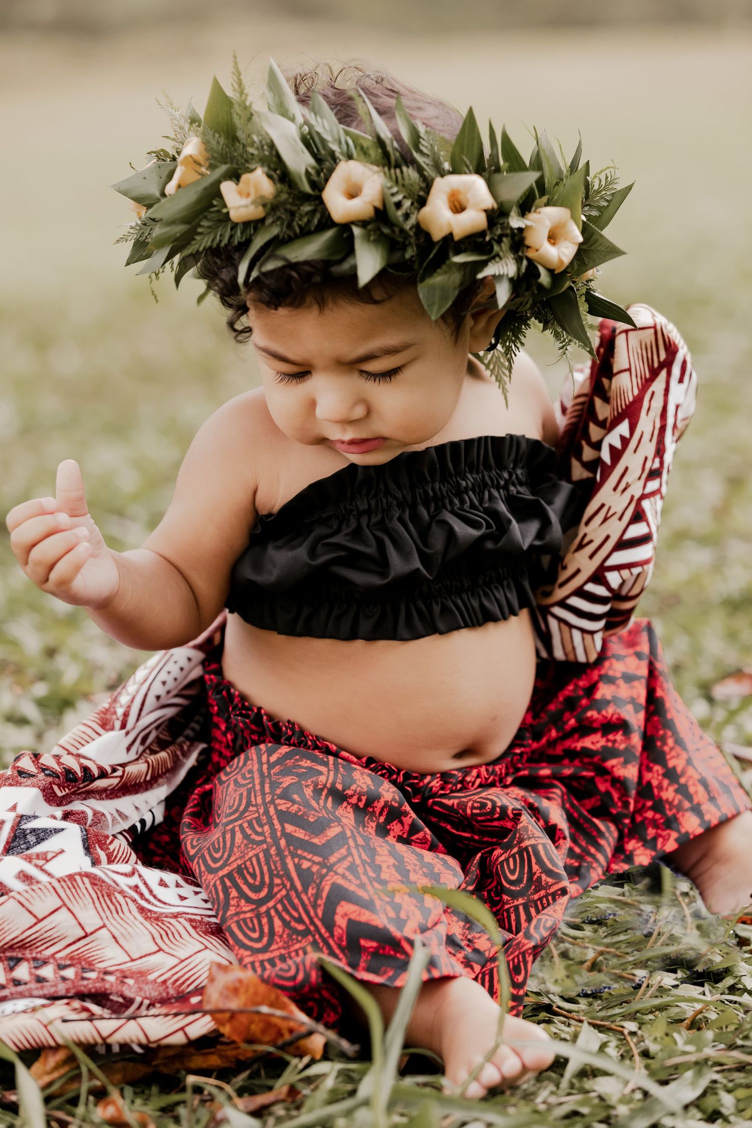 A baby wearing a floral crown and traditional Hawaiian outfit sits in the grass.