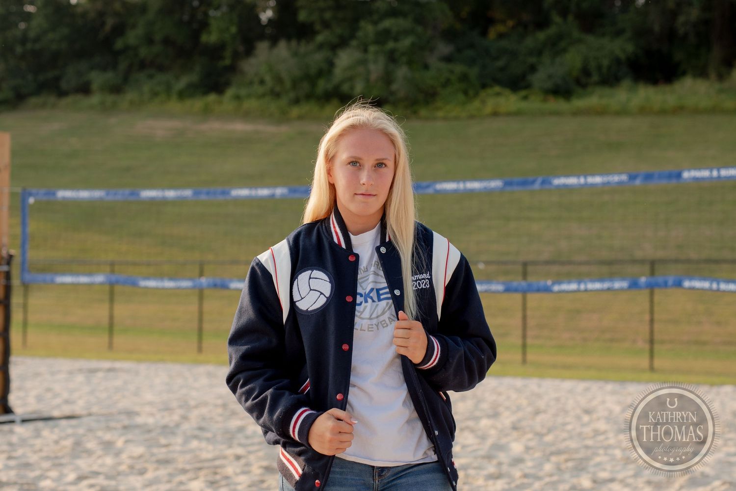 Athlete in letterman jacket standing on volleyball court with trees in background.