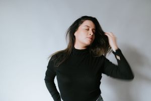 Dramatic portrait of person in black turtleneck against white wall with flowing dark hair.