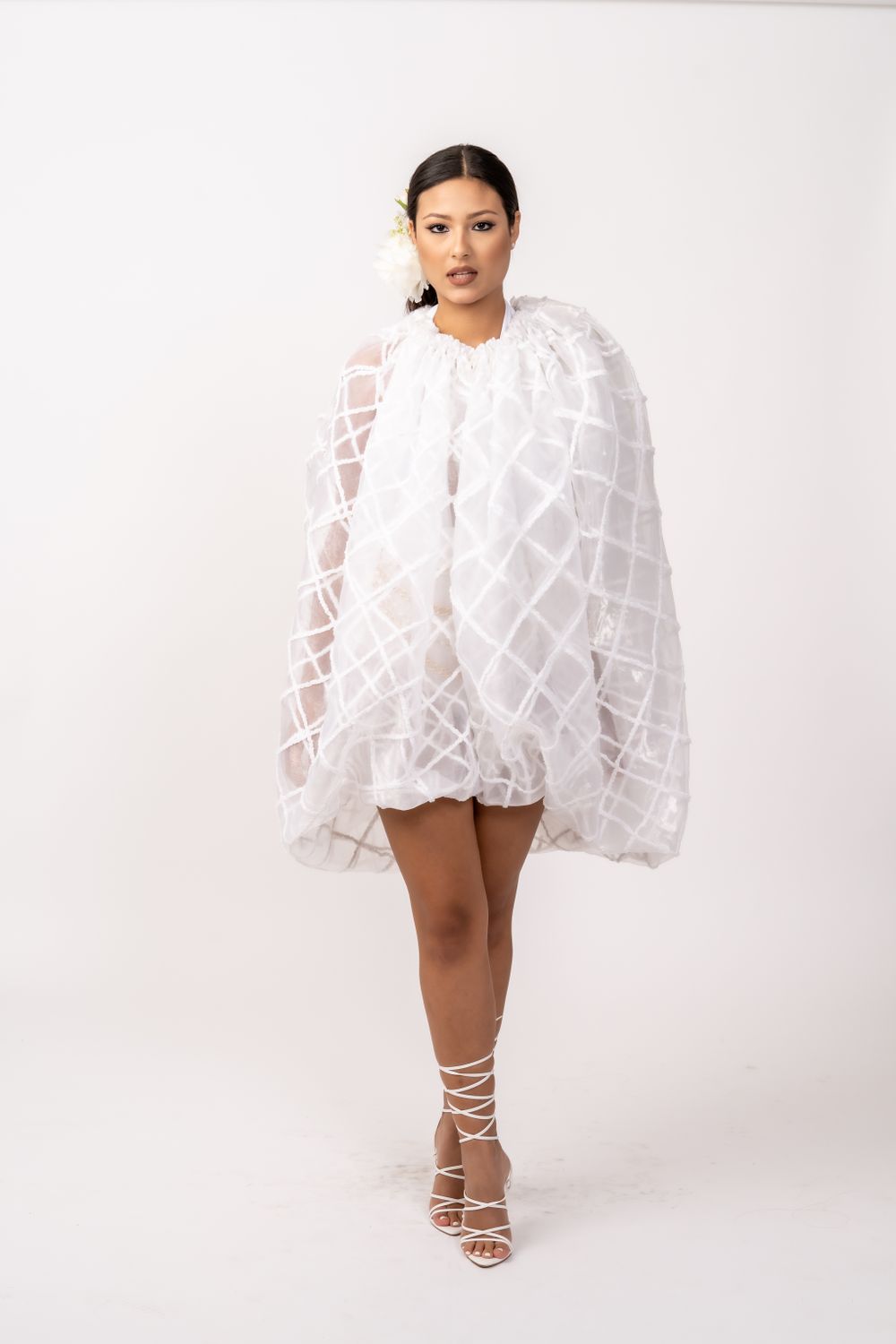 Fashion model wearing white textured cape dress with strappy heels against white background.