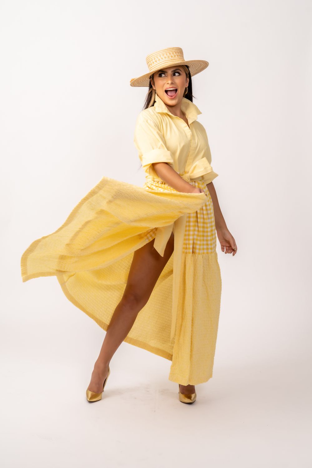 Dynamic pose in flowing yellow summer dress and straw hat against white background.