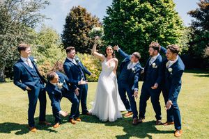 Wedding party poses playfully on sunny lawn in navy suits and white dress.