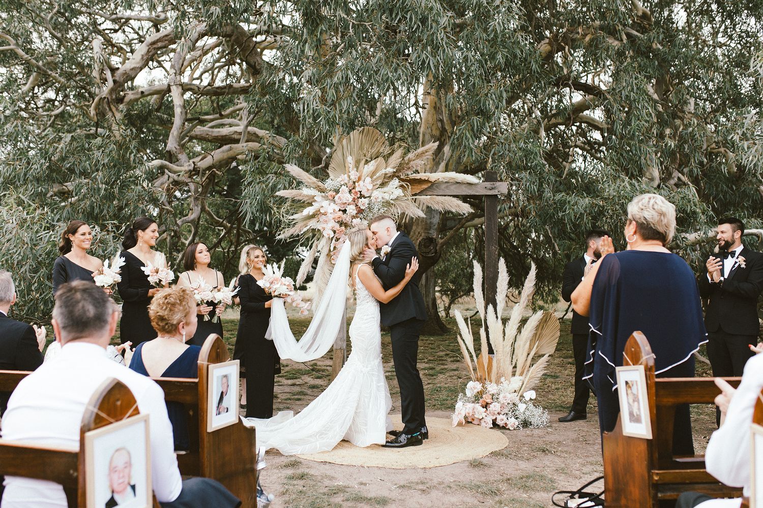 Outdoor garden wedding ceremony with wooden benches and eucalyptus trees creating natural setting.