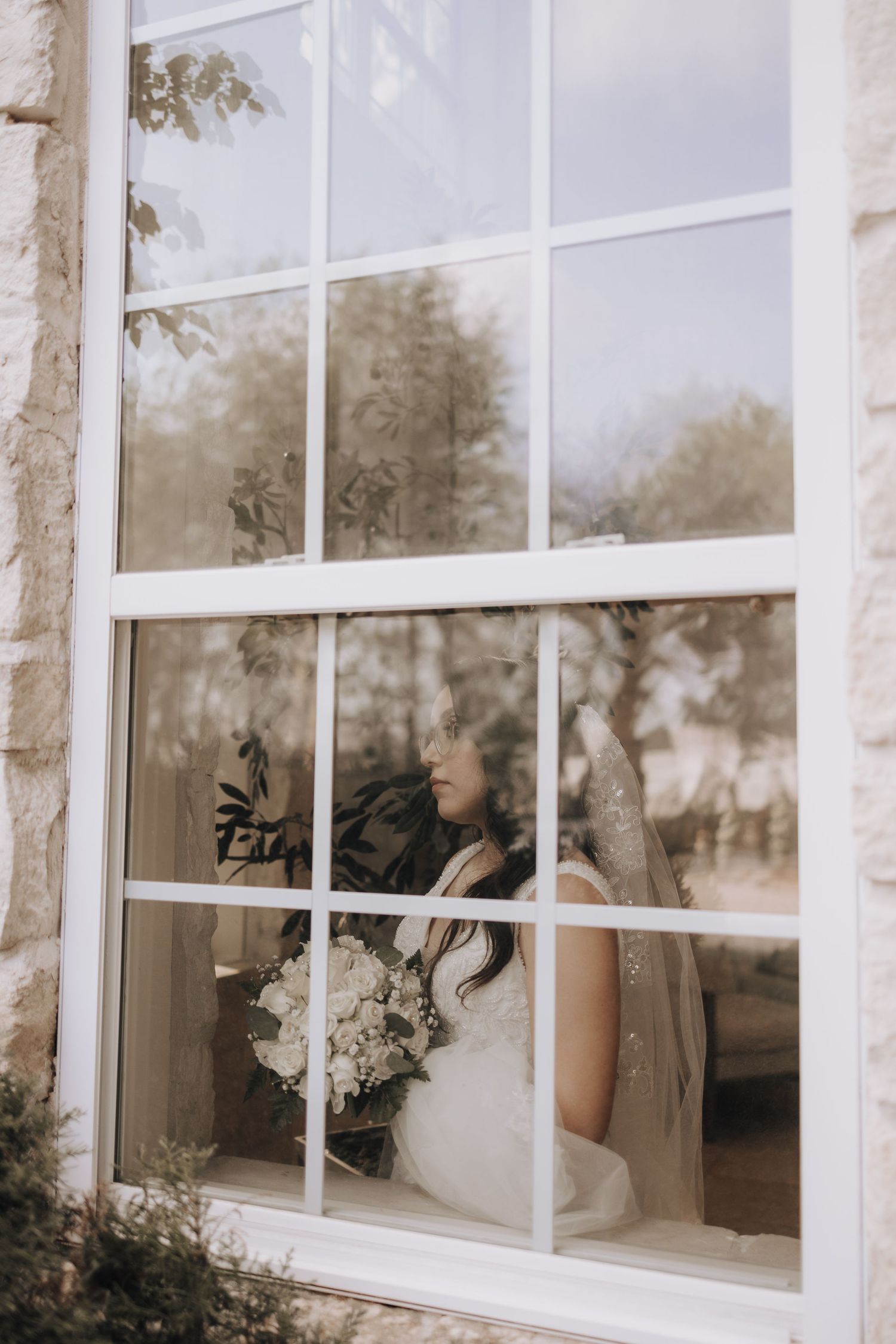 Through a window pane, a figure in white holding a bouquet is visible sitting indoors with soft natural lighting.