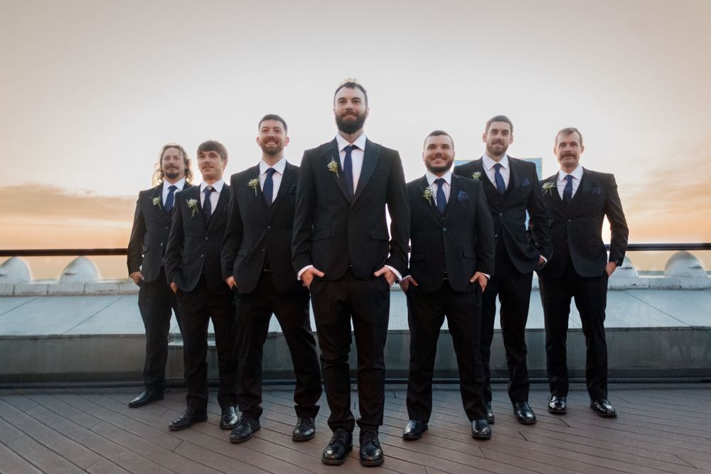 Groomsmen in black suits pose for a formal photo on a rooftop terrace.
