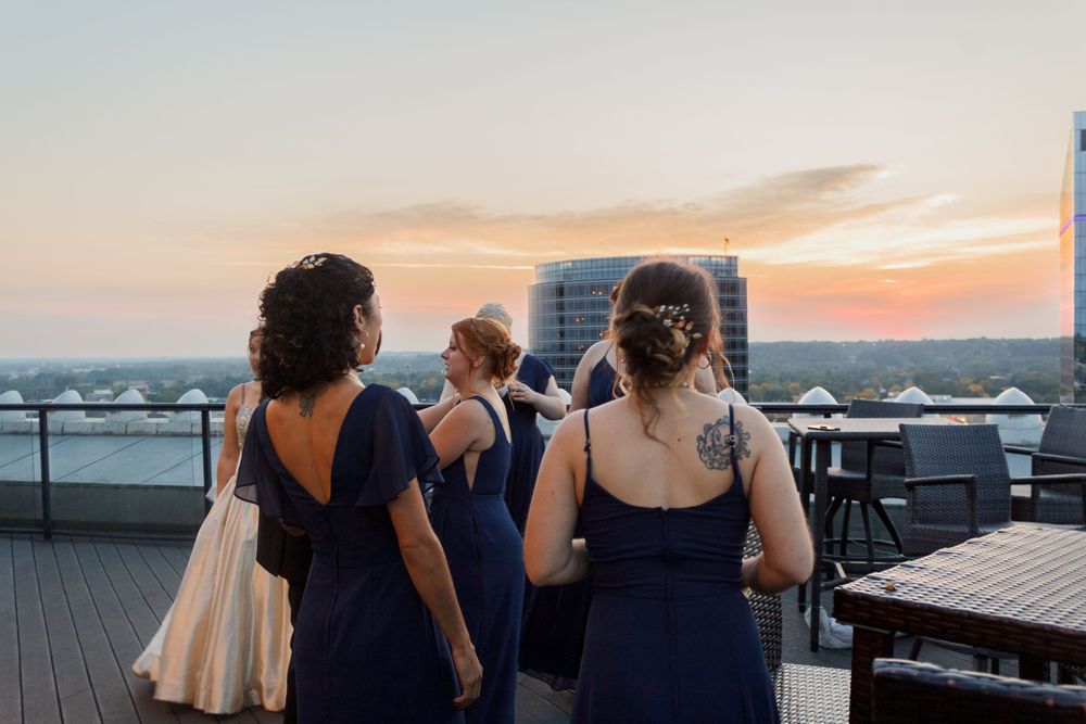 Three bridesmaids in navy dresses stand on a rooftop terrace at sunset.