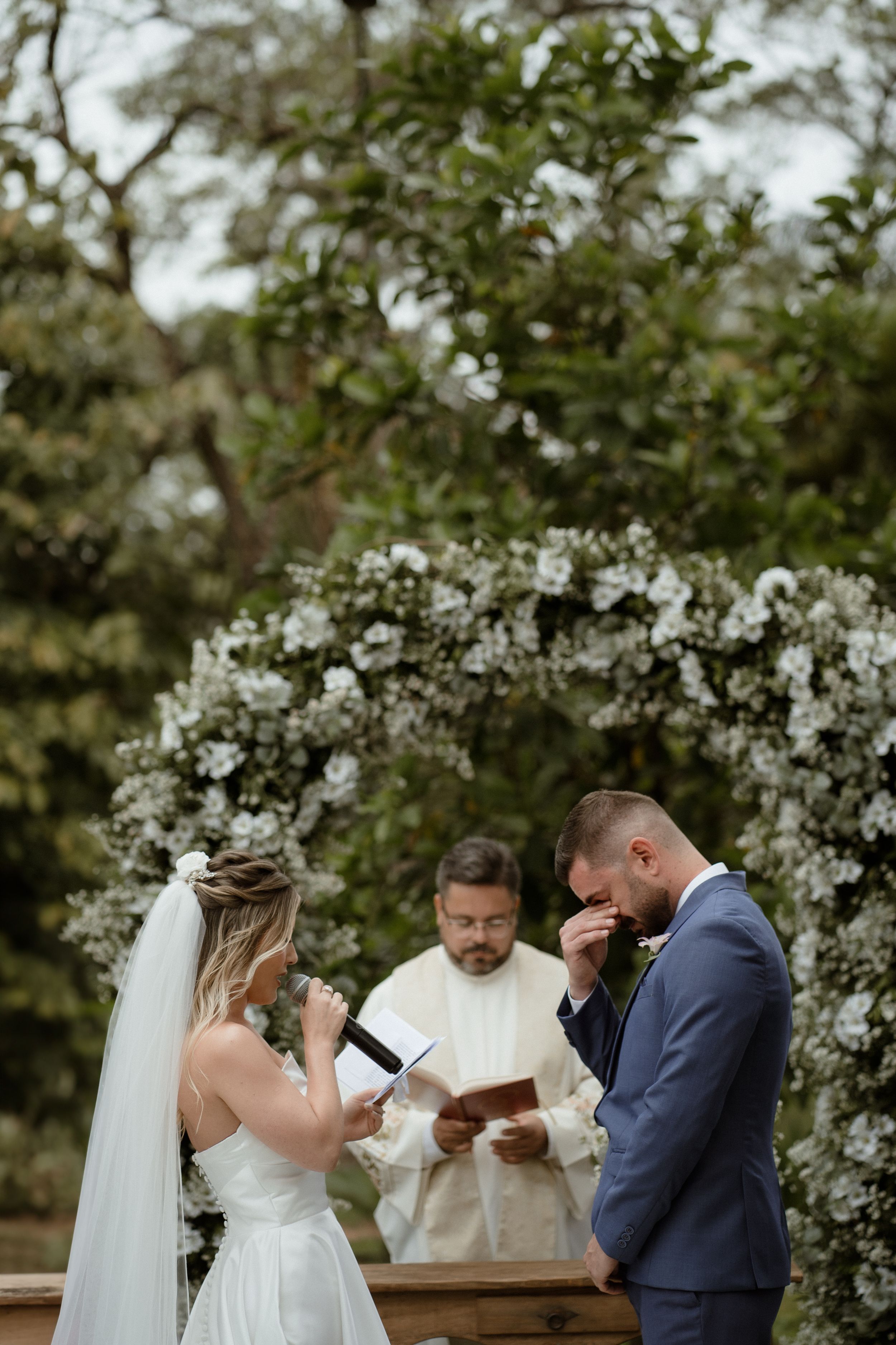 A wedding ceremony taking place outdoors with white floral decorations in the background.