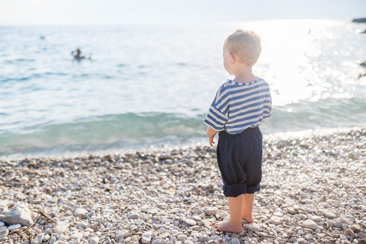 Young child in striped shirt and shorts standing on a rocky beach near the ocean.