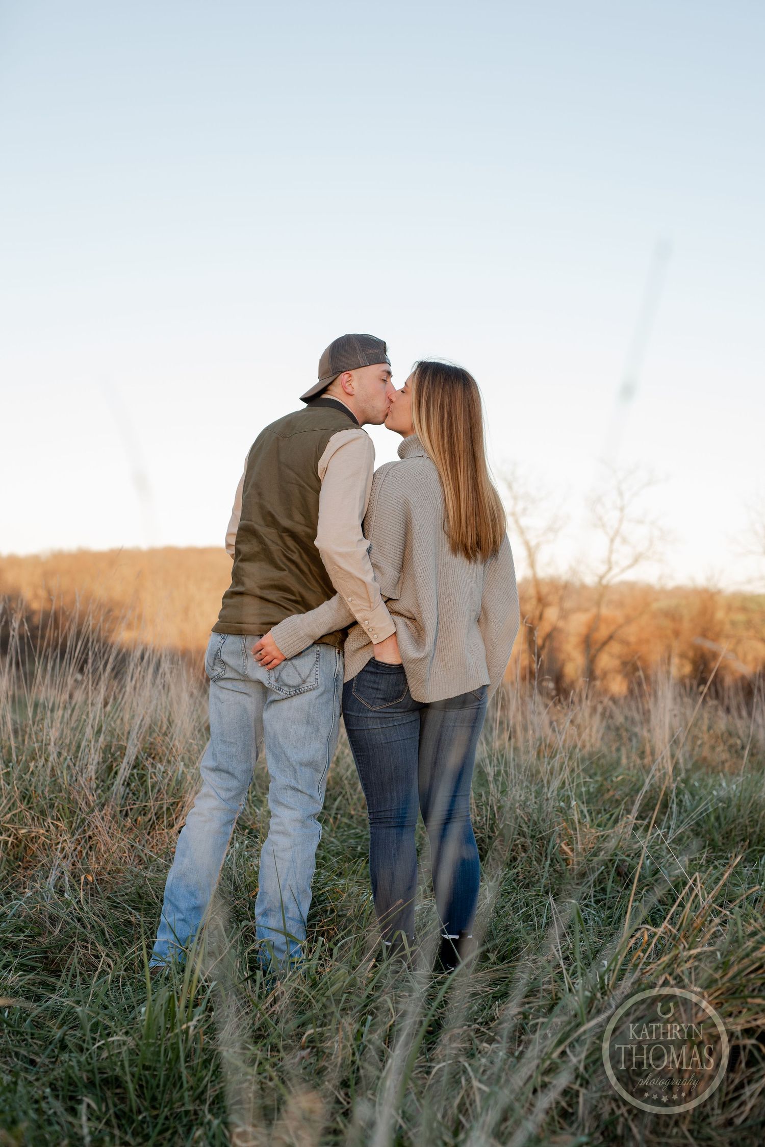 A romantic couple embraces while standing in tall grass during golden hour in a rural field.