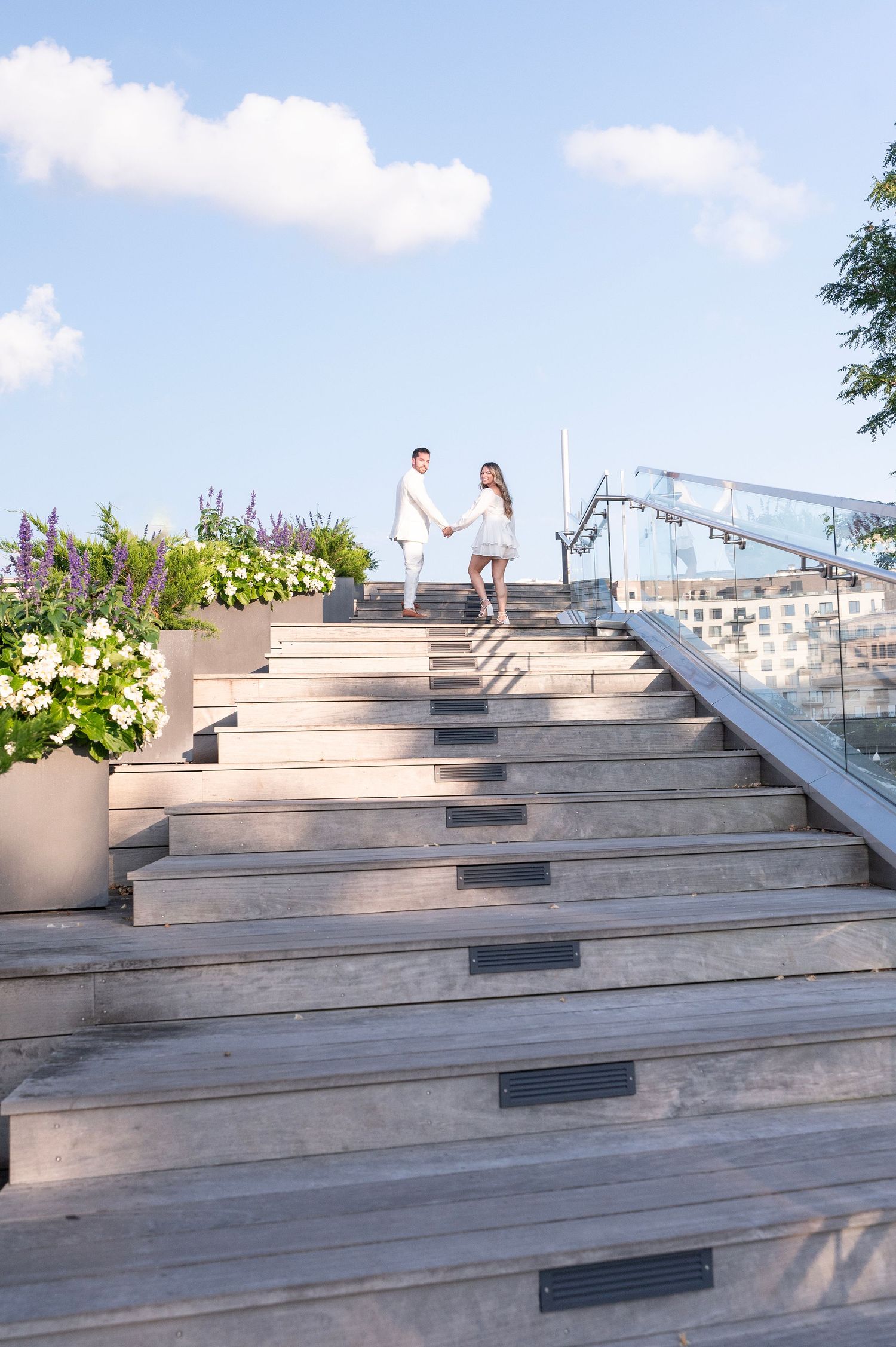 Couple descending wooden stairs with potted flowers and silver railing on a bright summer day.