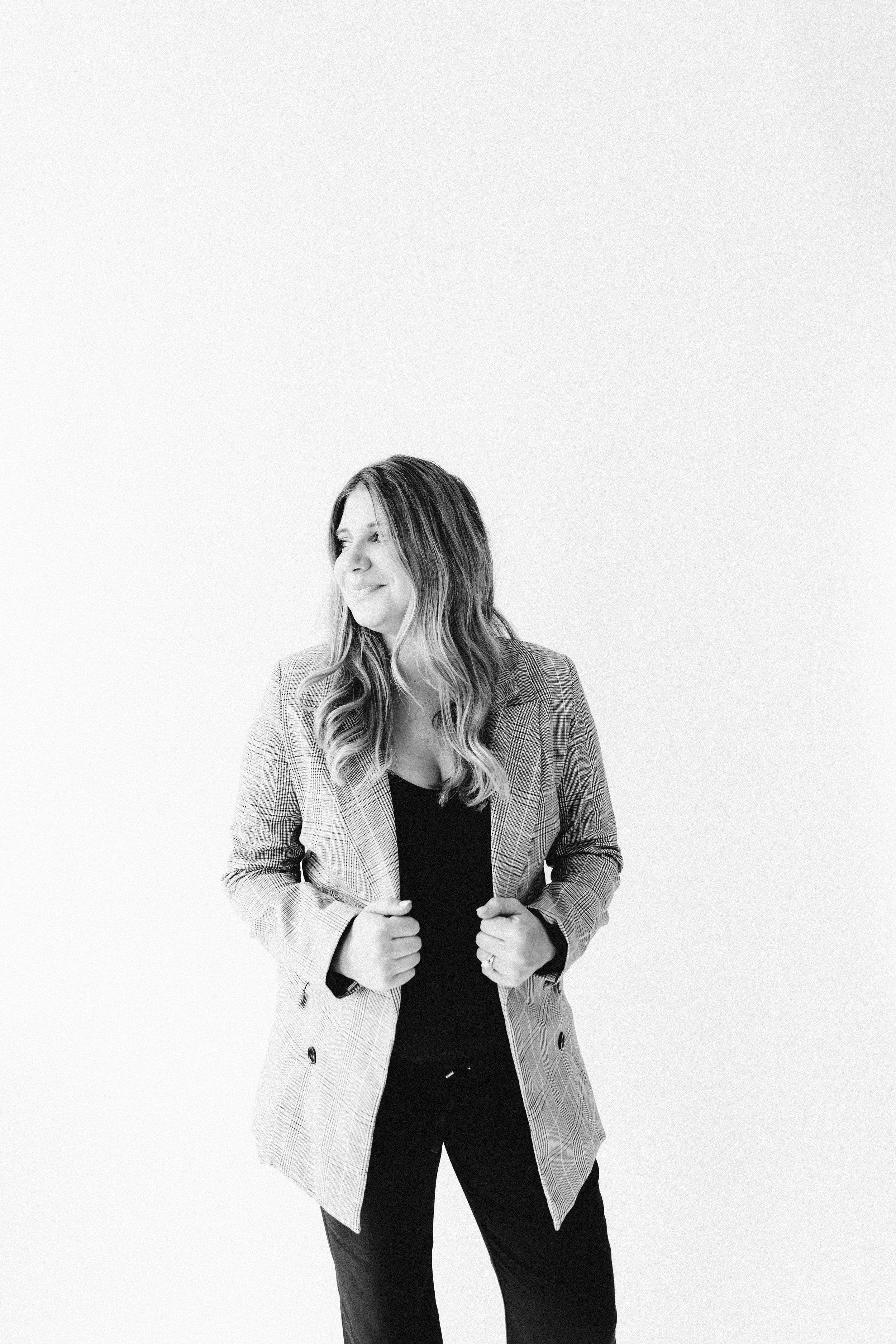 Woman in black outfit and gray blazer poses against white background in studio portrait.