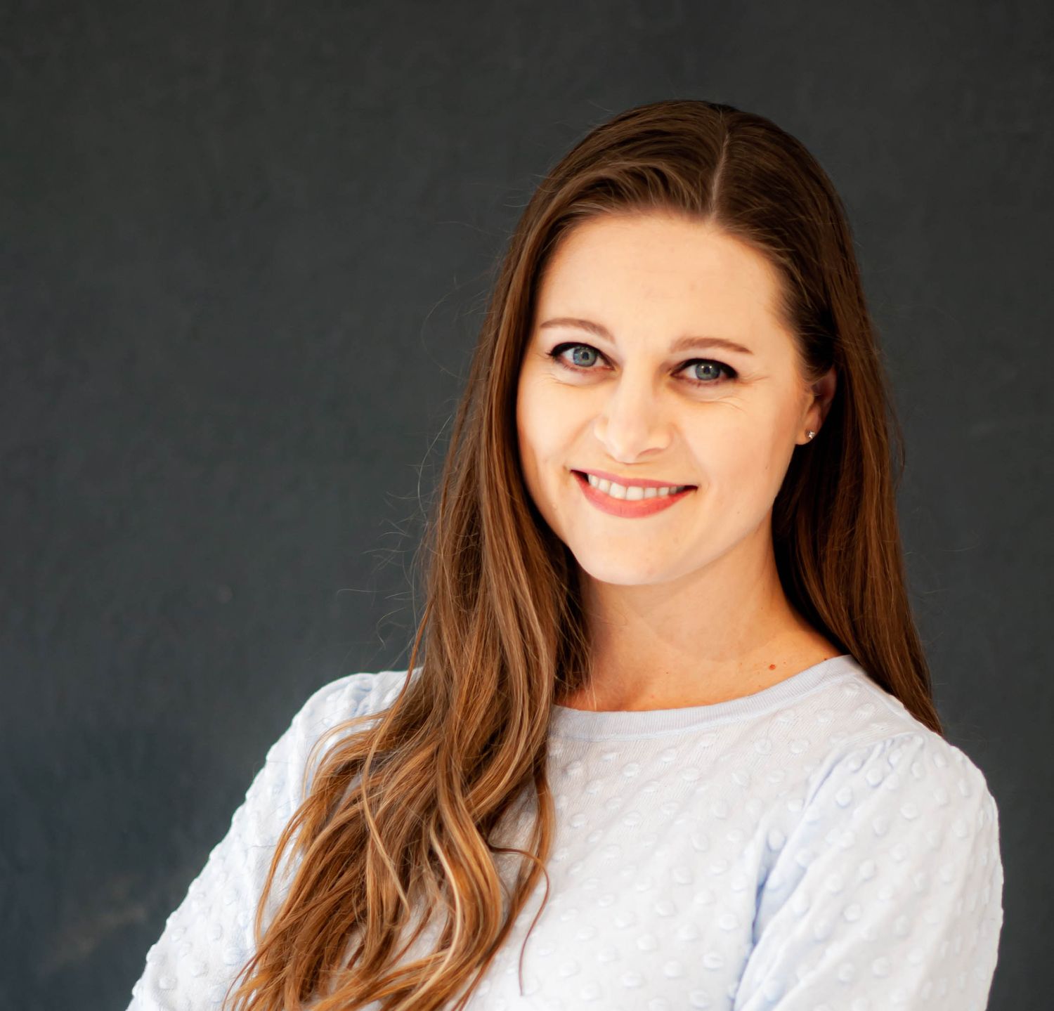 Professional headshot of a smiling young woman with long brown hair wearing a light blue sweater on grey background.