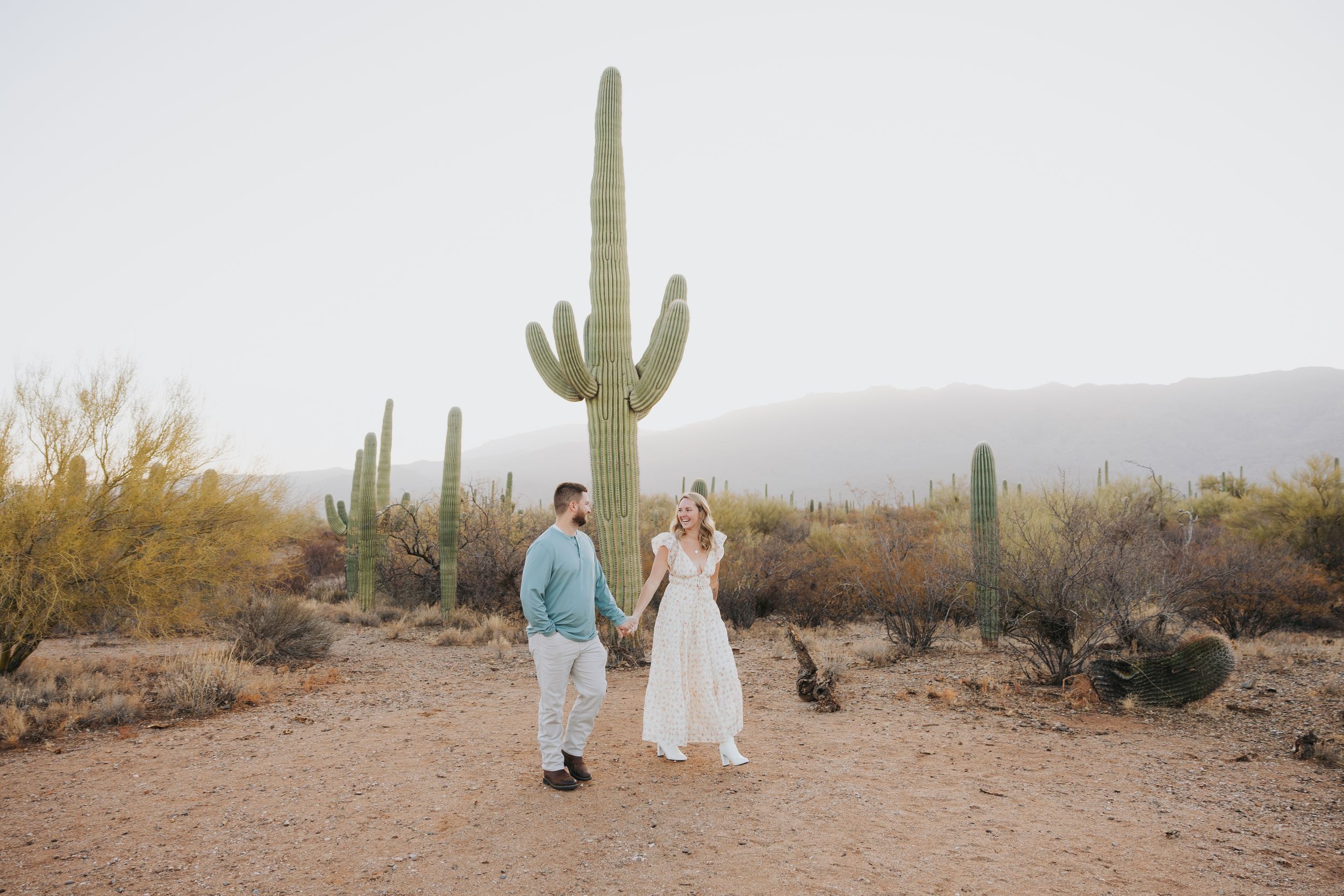 A couple poses near towering saguaro cacti in a desert landscape at sunset.