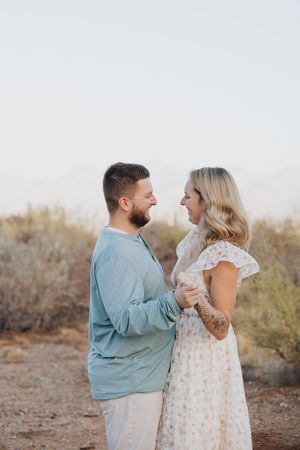 A romantic couple embraces in a desert landscape at sunset wearing coordinated blue and white outfits.