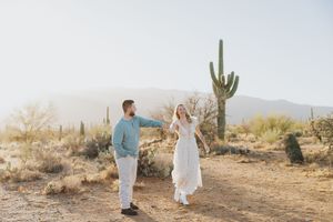 A couple holds hands while dancing along a desert path surrounded by saguaro cacti.
