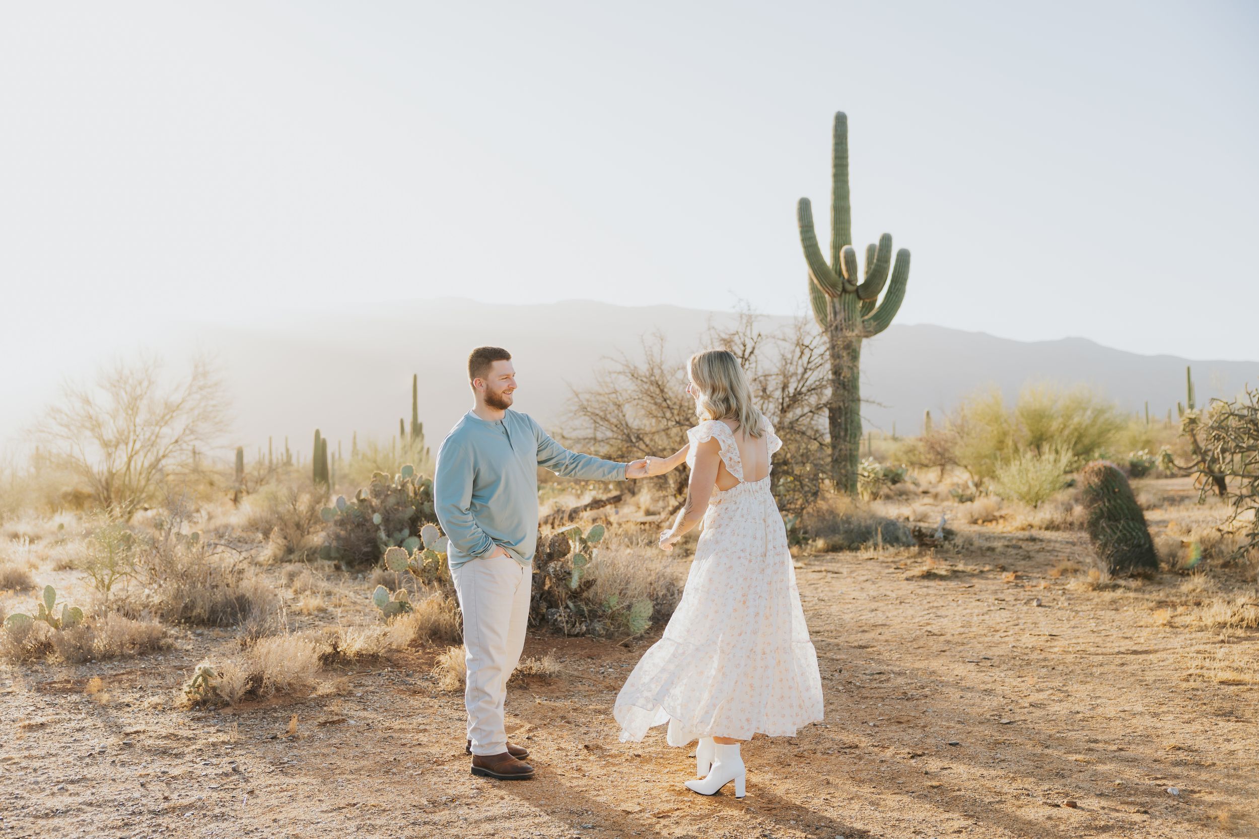A couple in elegant attire poses in a desert landscape with towering saguaro cacti at sunset.