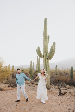 A couple holds hands while walking through a scenic desert landscape with giant saguaro cacti.