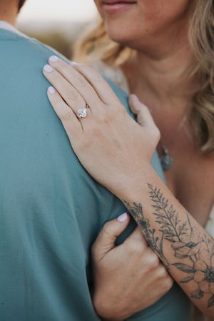 Close-up of hands showing floral tattoo and engagement ring against a turquoise shirt.