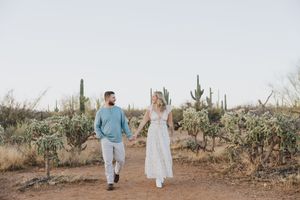 A couple walks hand in hand through a desert landscape dotted with saguaro cacti.