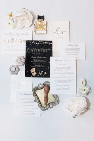 Wedding invitation suite with black and white stationery, floral accents, perfume bottle and vintage mirror on light surface.