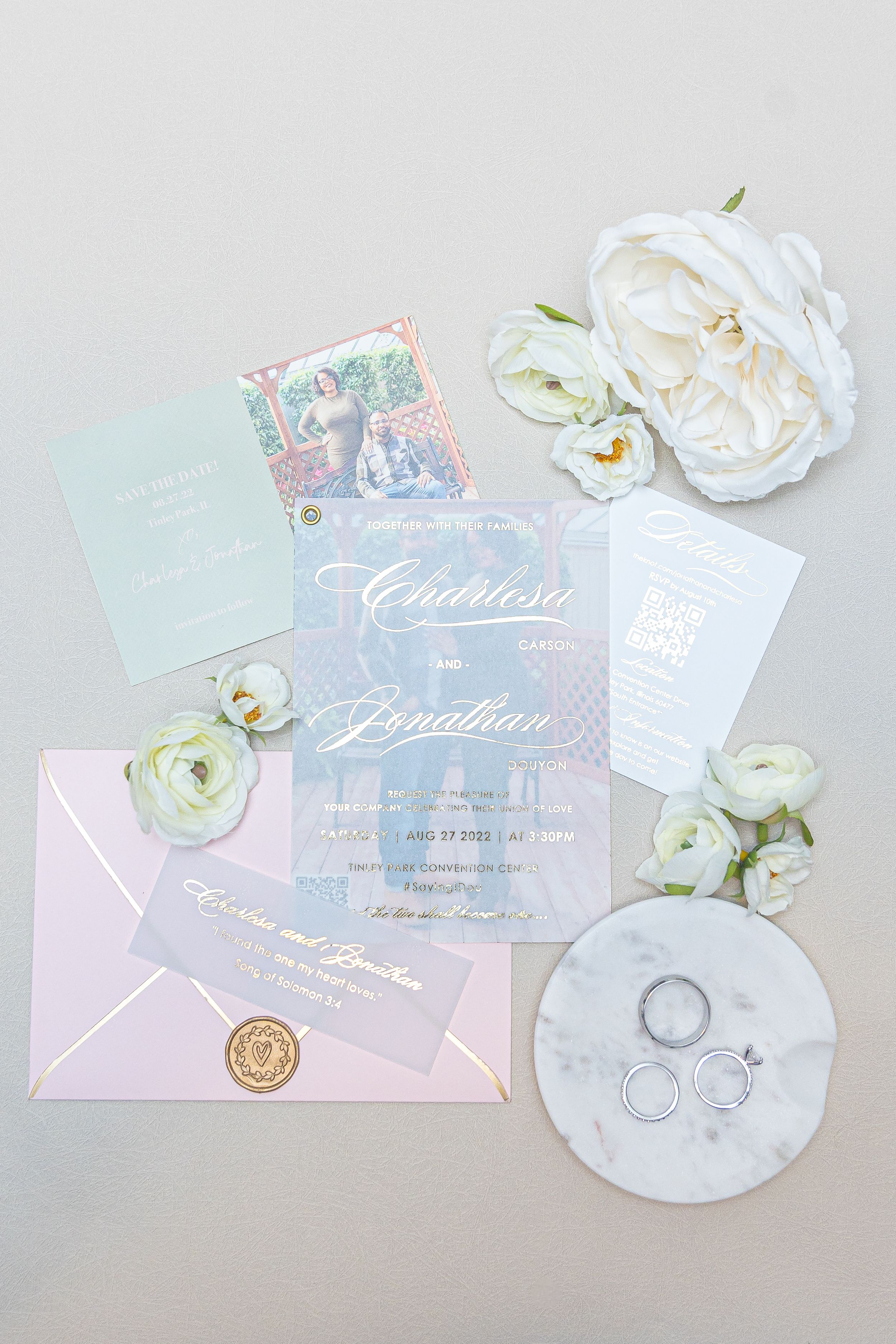 Wedding invitation suite with pink and blue colors displayed with rings, white flowers and gold wax seal on neutral background.