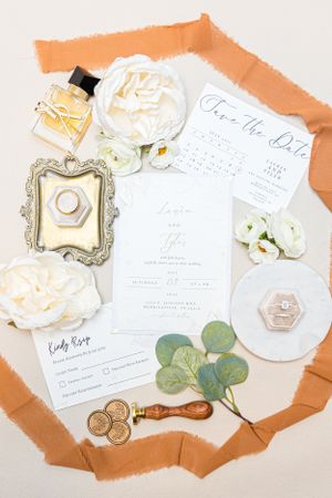 Wedding stationery flatlay with white roses, perfume bottle, vintage frame, wax seals and eucalyptus on peach background.