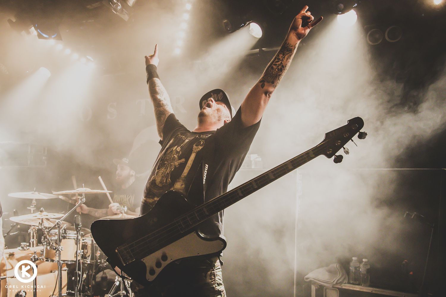 A bassist performs energetically on stage with dramatic backlighting and fog effects during a concert performance.