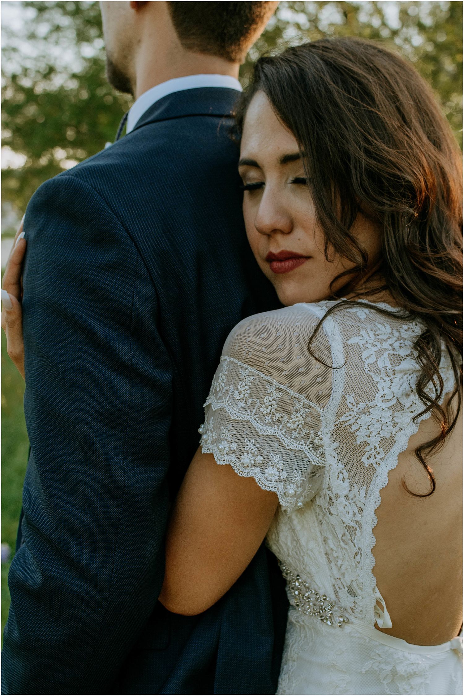 Close-up of bride's lace detailed dress back against groom's navy suit in natural setting.