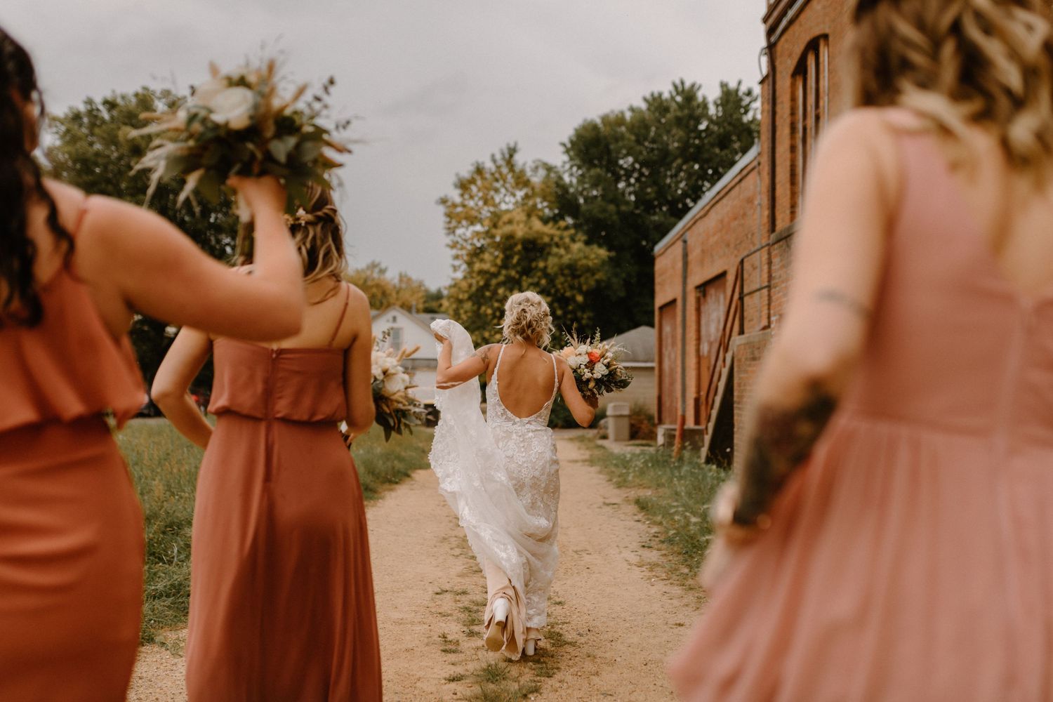 A bride runs along a dirt path while attendants in coral dresses watch.