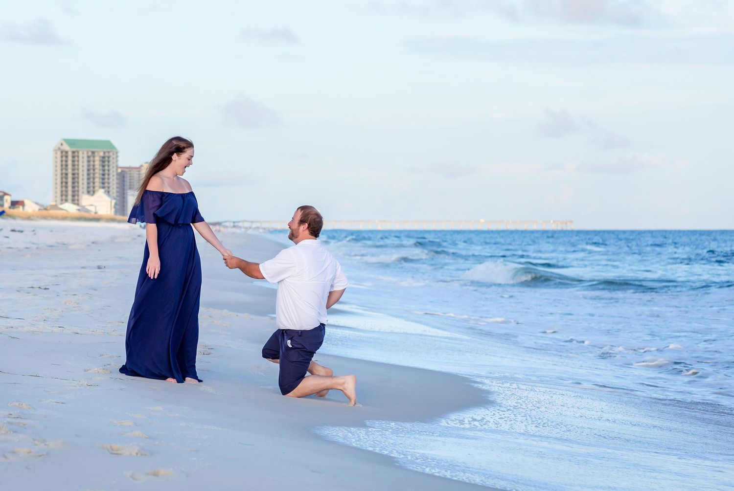 Romantic beach proposal scene with waves crashing and coastal buildings in background.