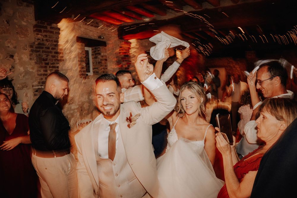Wedding reception guests celebrate and dance together in a dimly lit rustic venue with brick walls and red lighting.