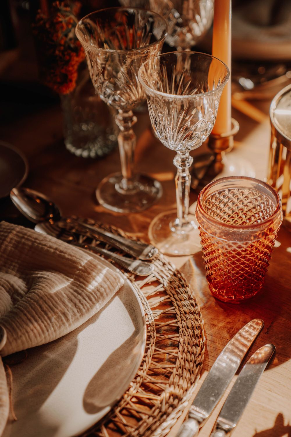 Elegant vintage place setting with cut crystal glassware and woven charger plates on a rustic wooden table in warm lighting.