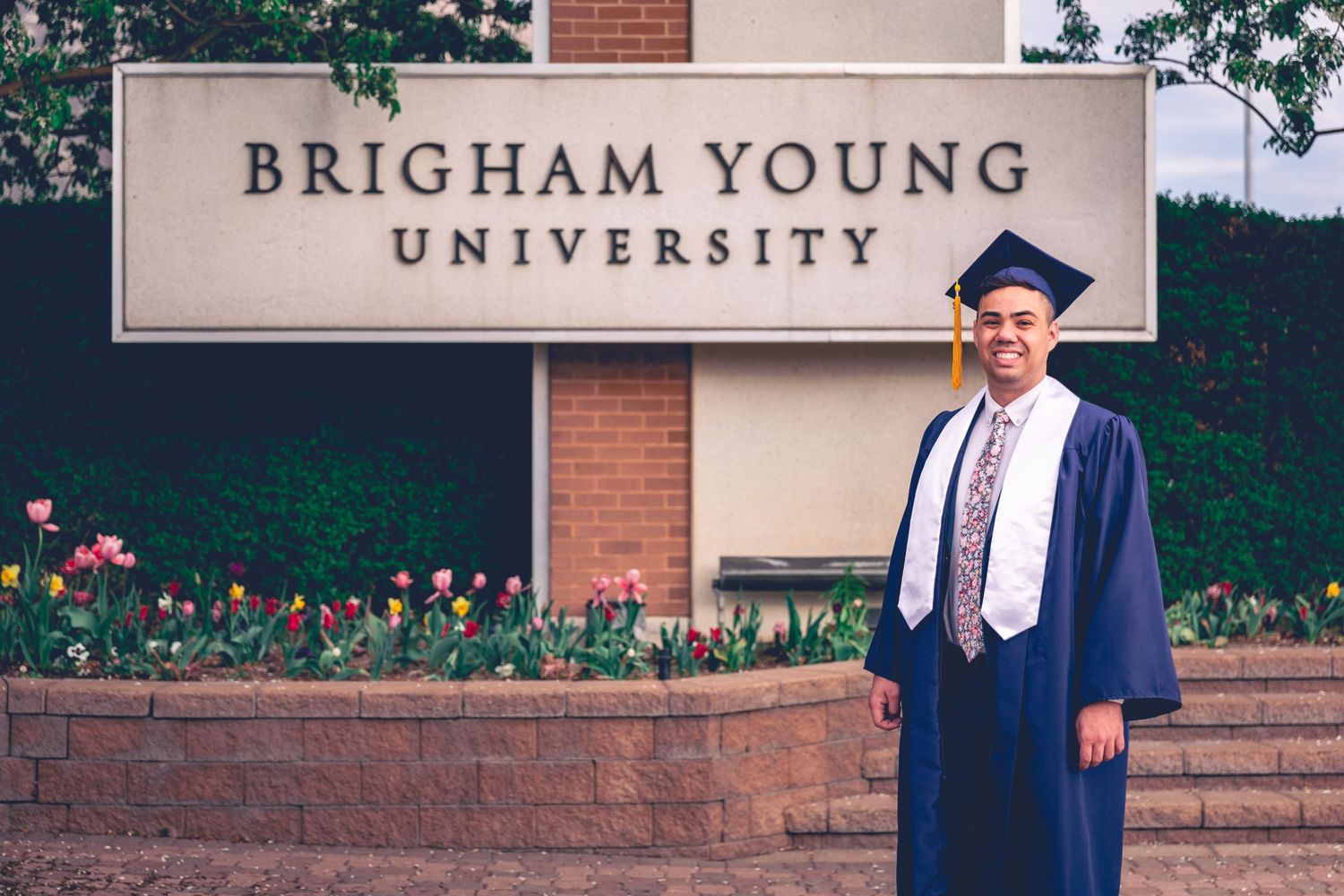 college graduate stands in front of brigham young university sign in cap and gown for graduation photoshoot