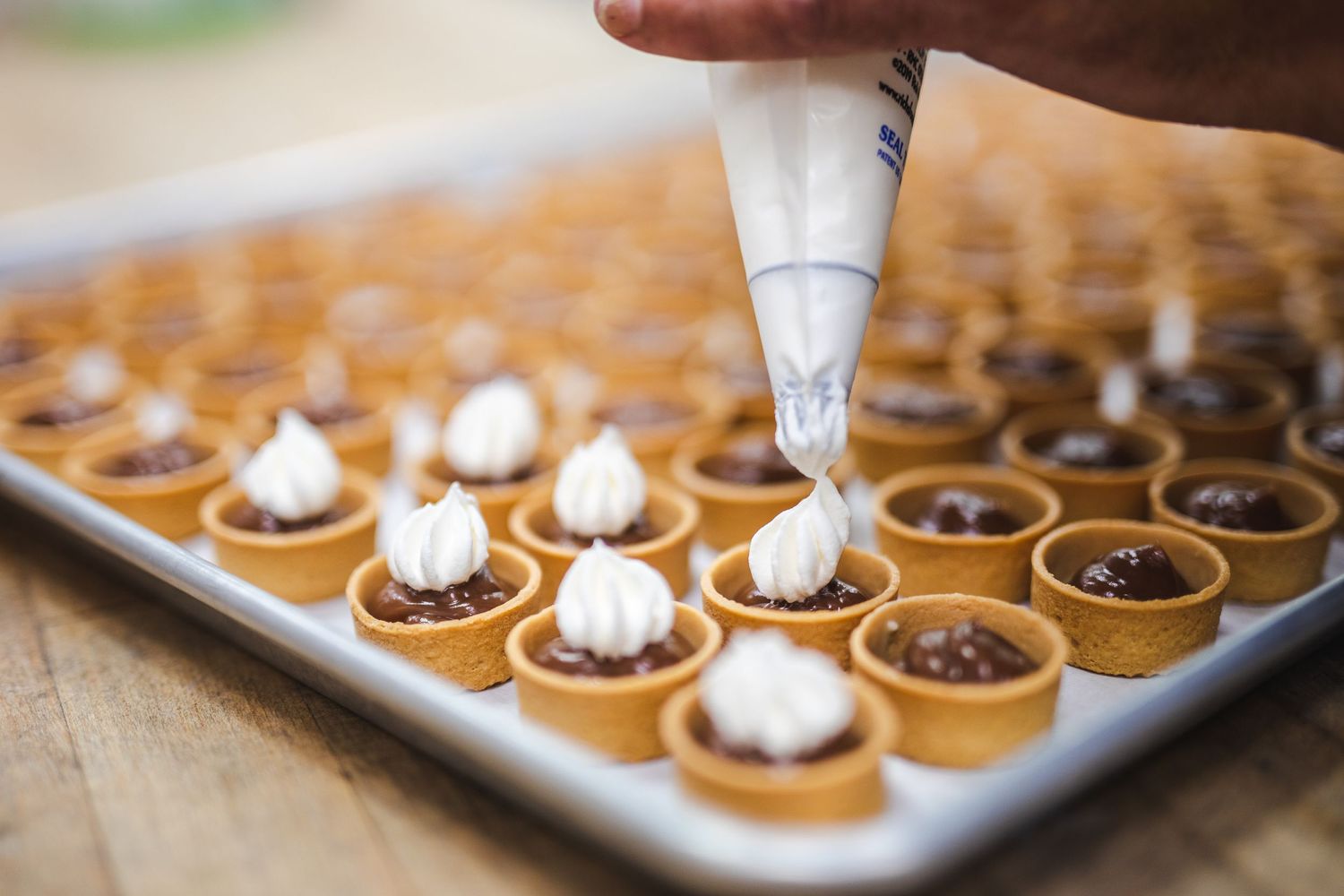 A pastry chef piping white frosting onto mini tarts arranged in rows on a baking sheet.