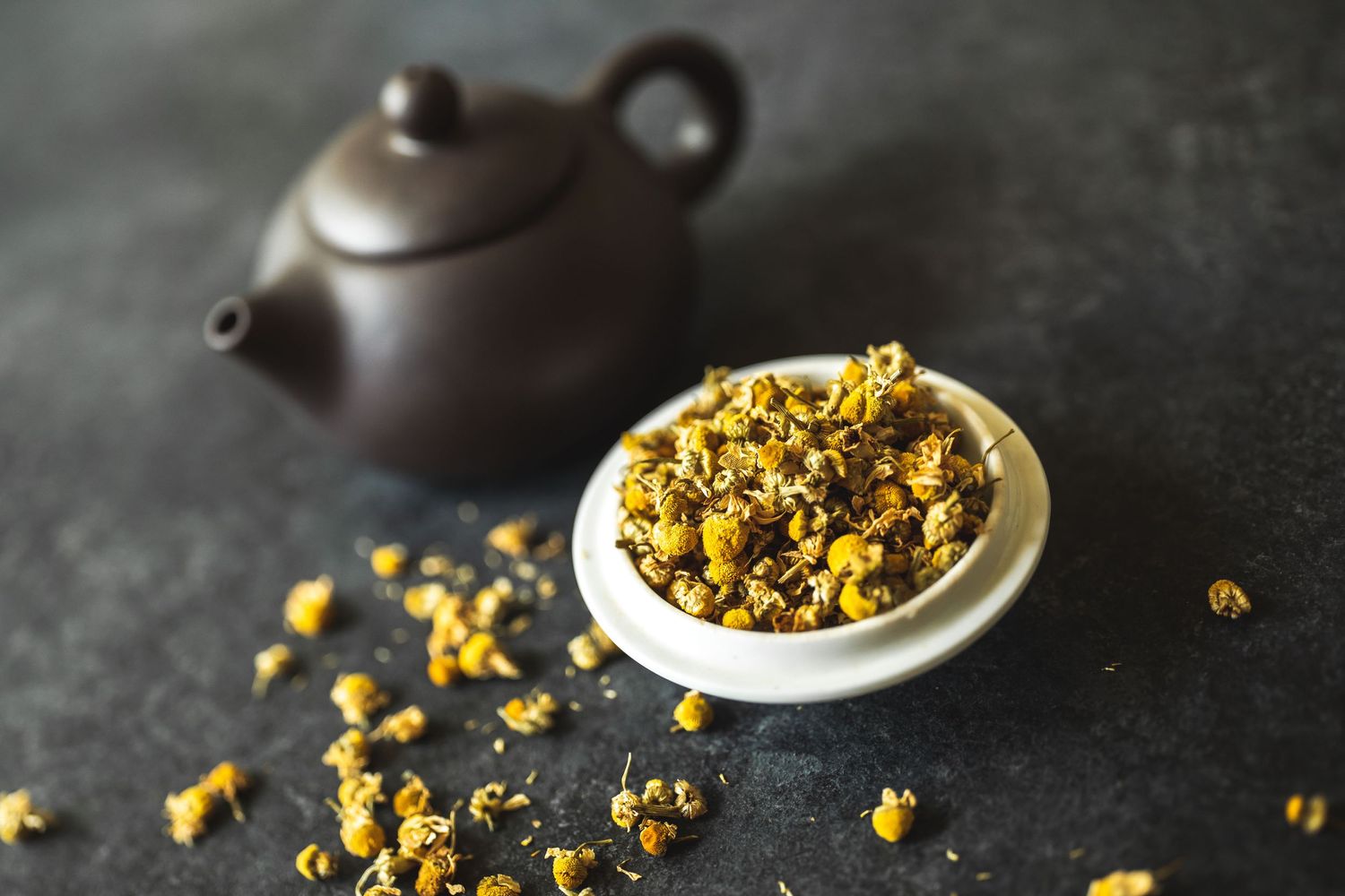 Brown teapot with dried chrysanthemum flowers in white dish on dark surface.
