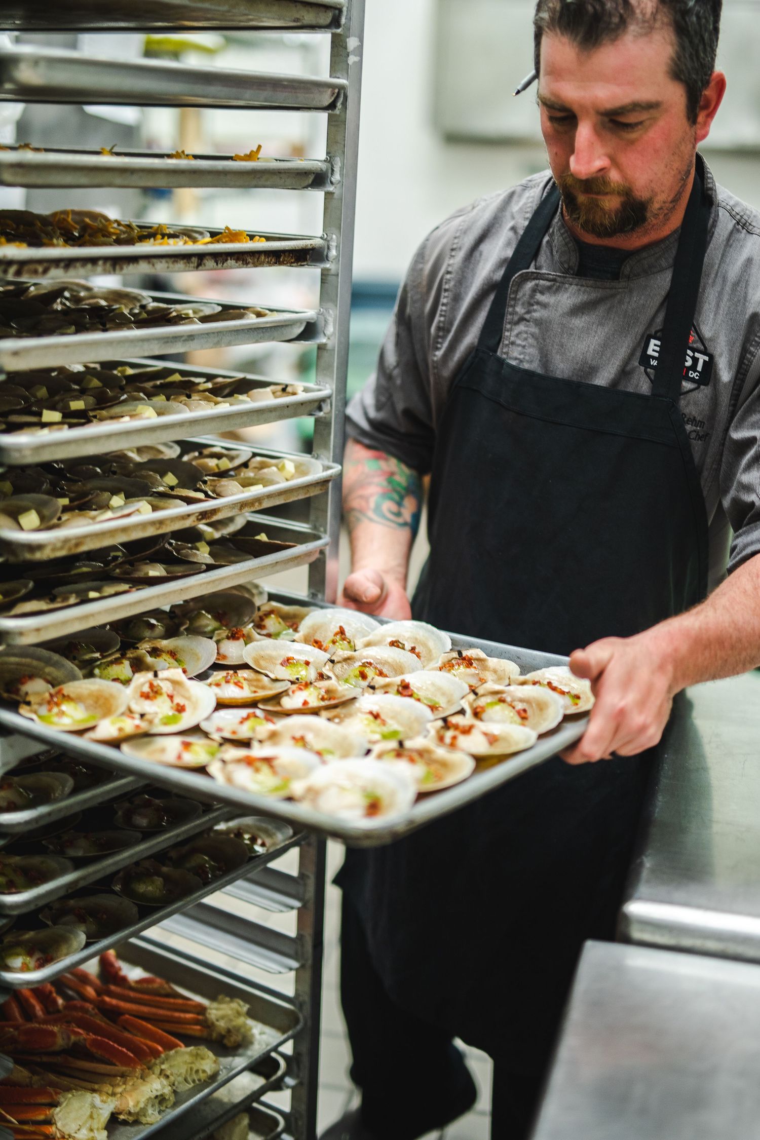Chef in commercial kitchen holding tray of prepared food near stacked sheet pans.