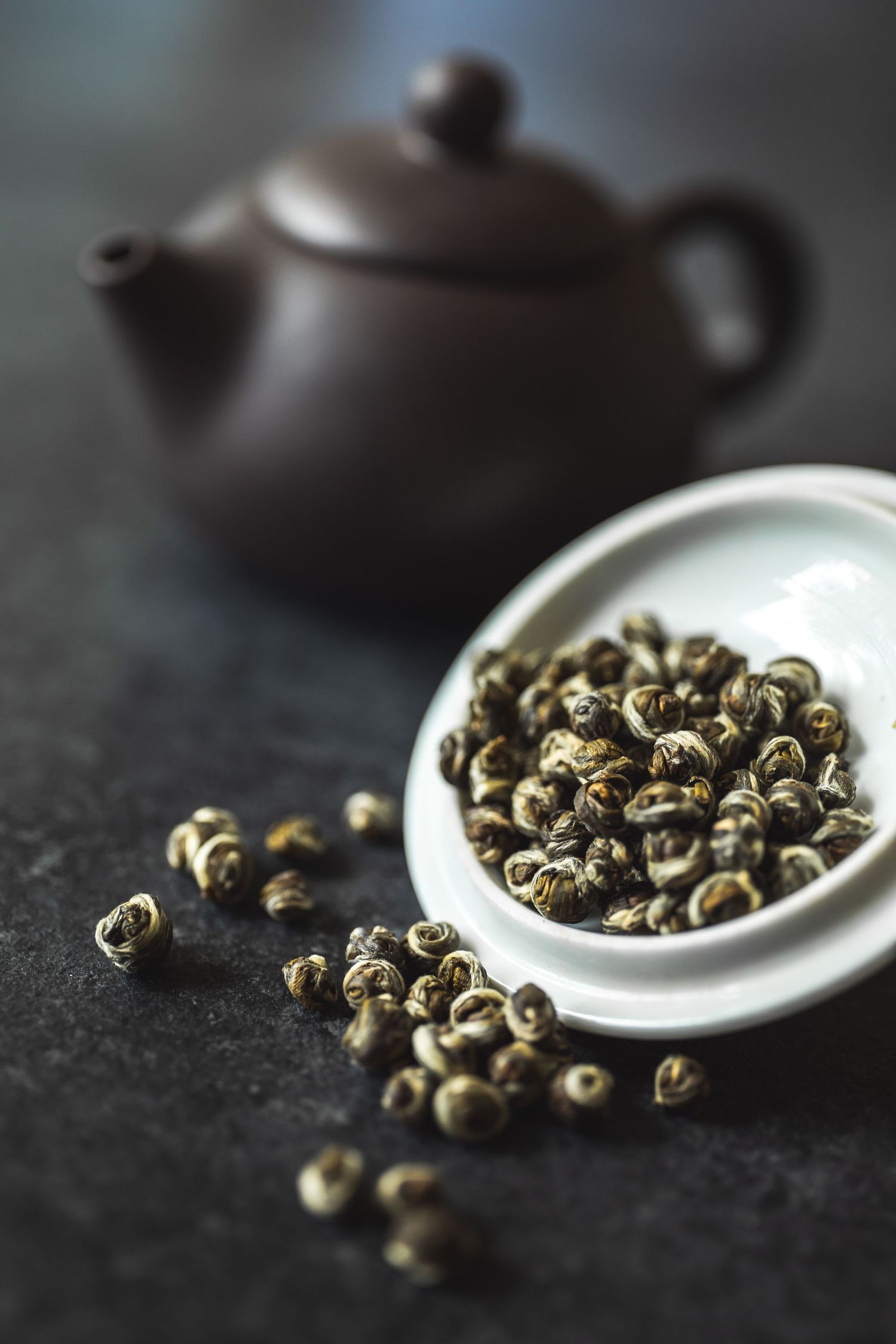 Traditional clay teapot with loose jasmine tea pearls on dark surface.