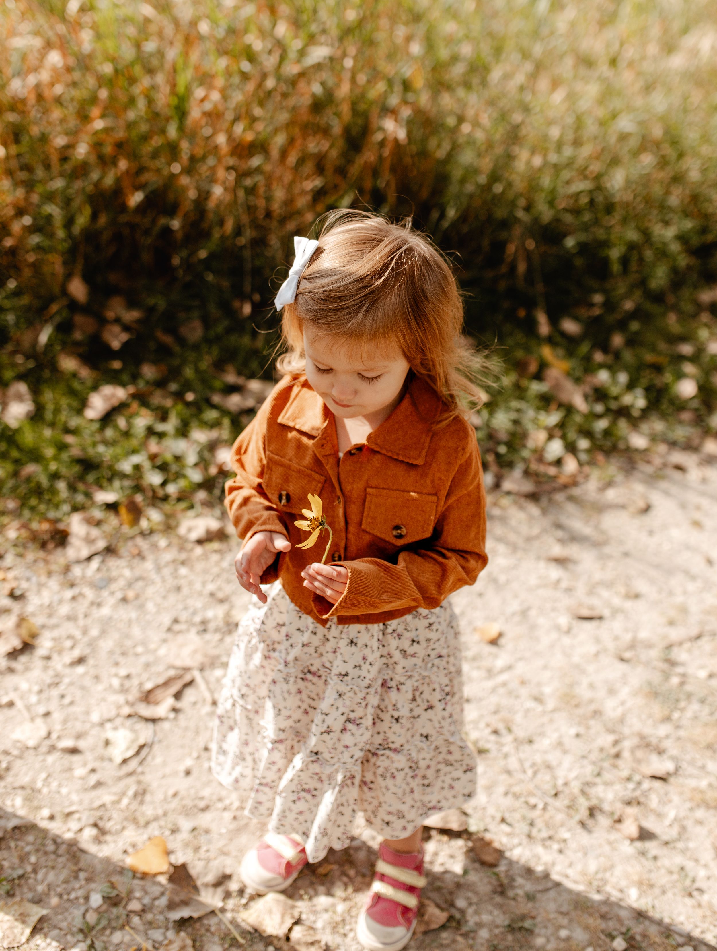 A small child in an orange jacket and floral skirt looking down at something.