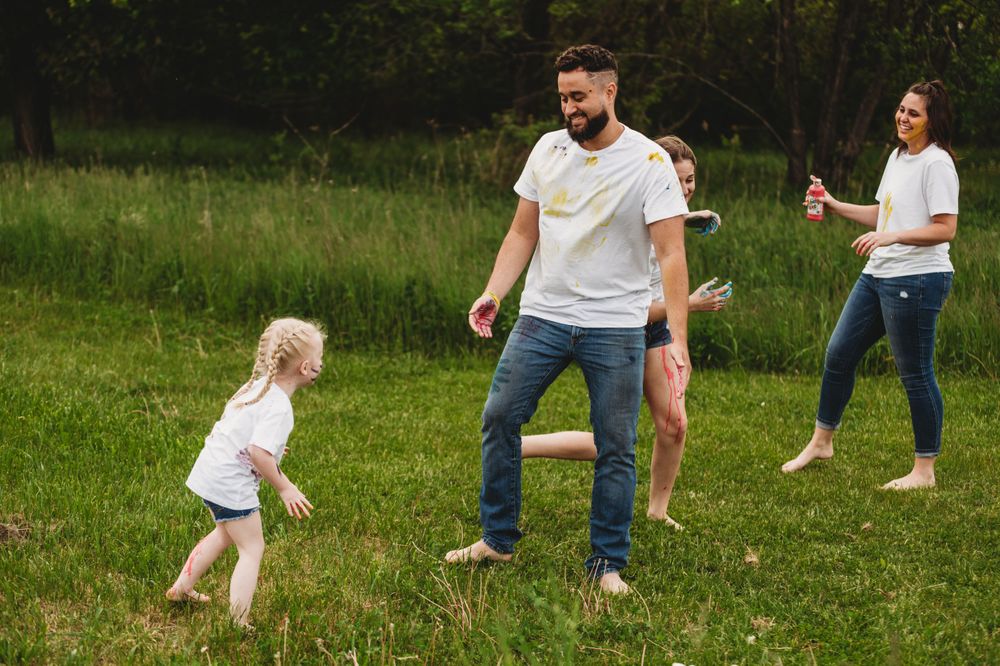 People playing with colorful paint in a grassy outdoor setting wearing white shirts and jeans.