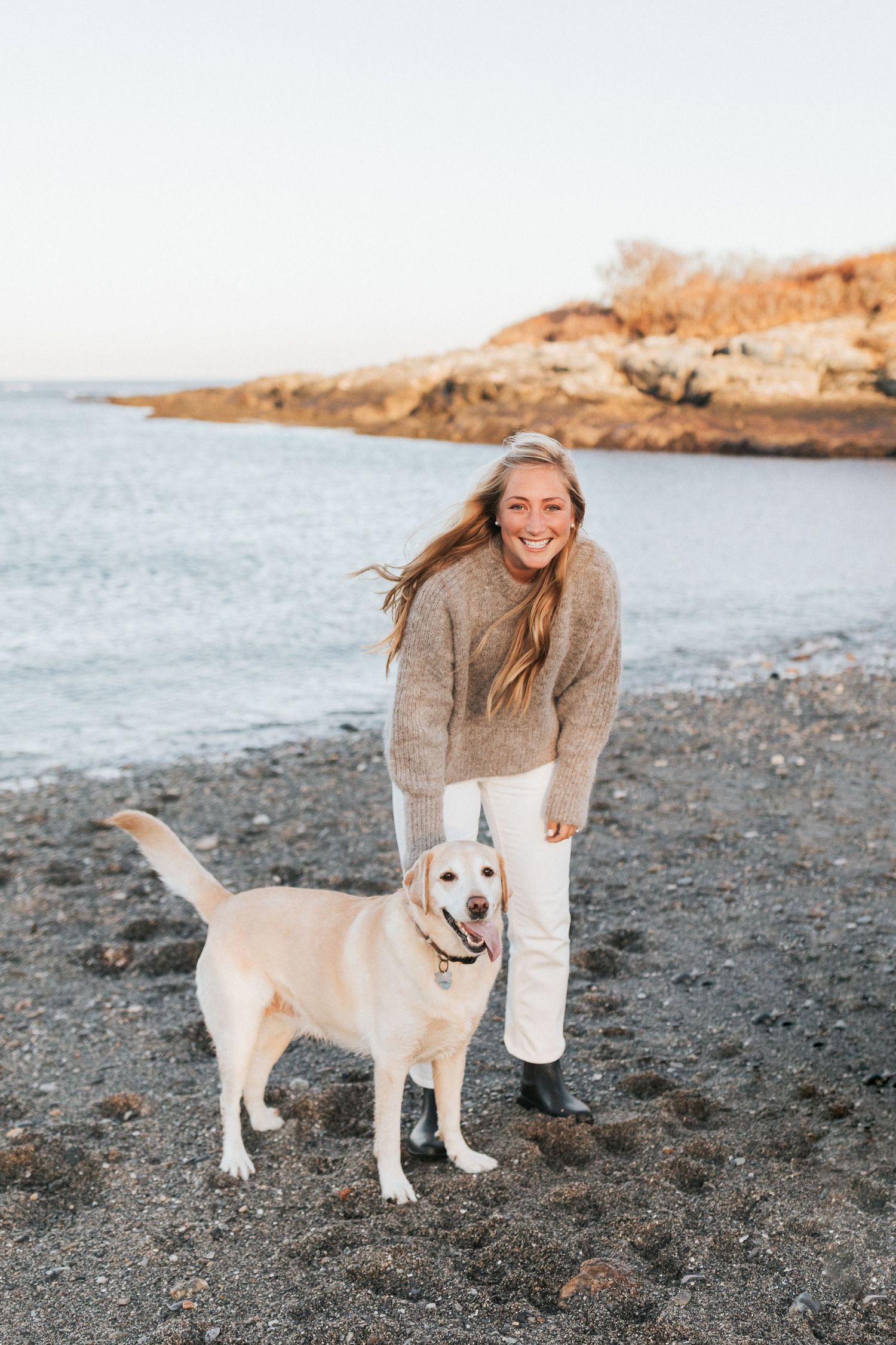 Person in beige sweater posing with friendly yellow Labrador dog on rocky beach.