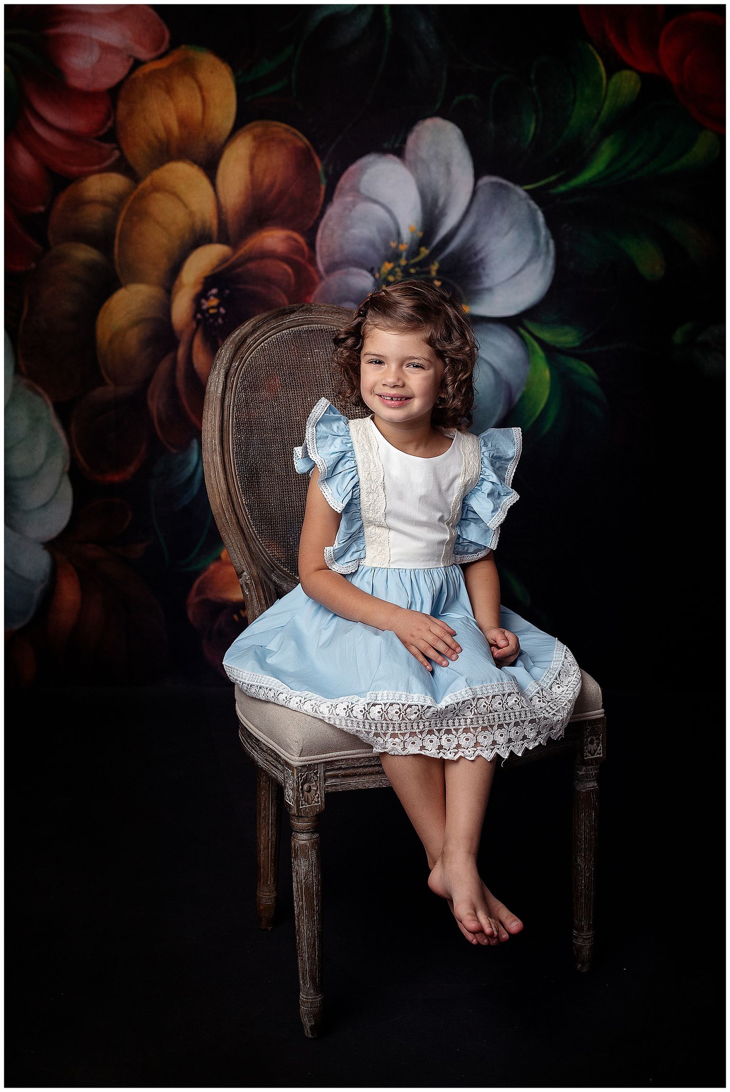 Valerie Kay Photography fine art child portrait featuring girl in blue apron dress sitting on chair w/ bold floral back
